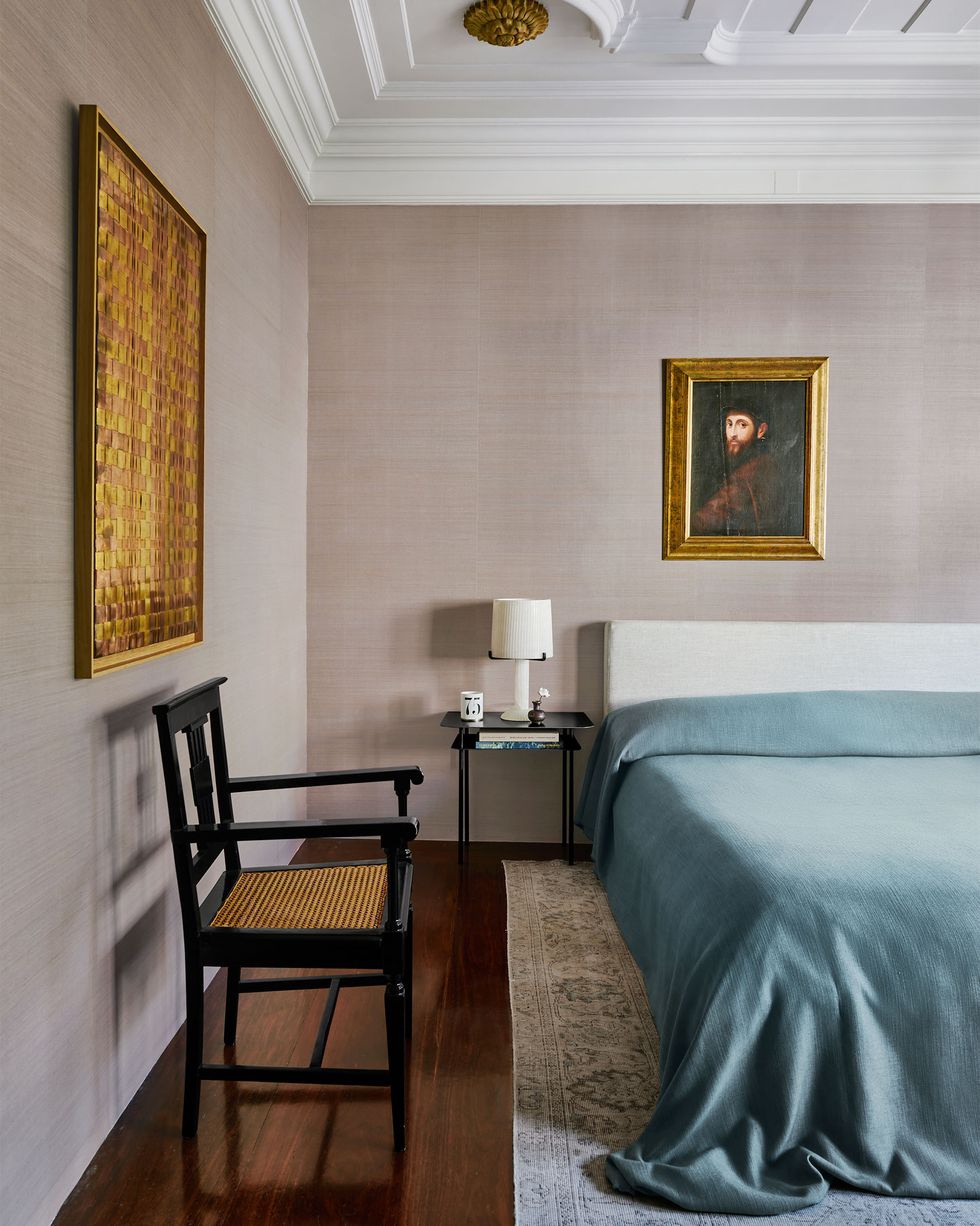 a bed with fabric headboard and a light blue linen cover, a black nightstand with a white lamp, a black lacquer wooden chair, a bleached kilim, and an old master painting and a woven metal artwork on walls