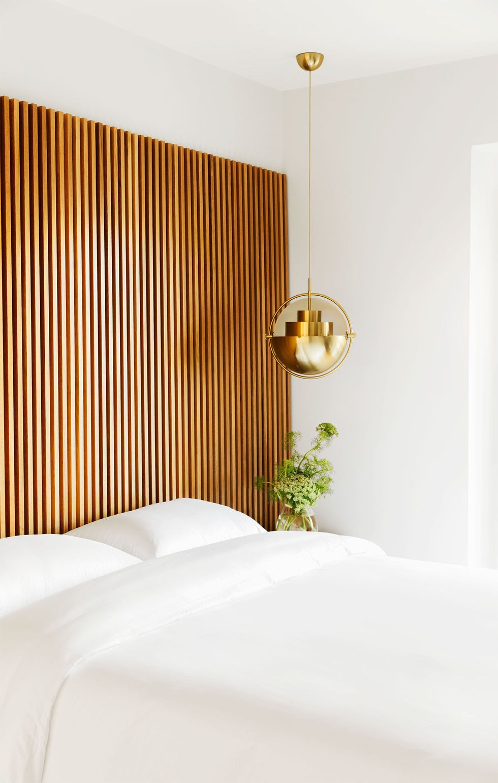 in the corner of a bedroom the walls and bedding are white, wooden slats line the wall behind the bed, beside it is a glass vase with light colored ferns and flowers, and above it is a spherical gold pendant