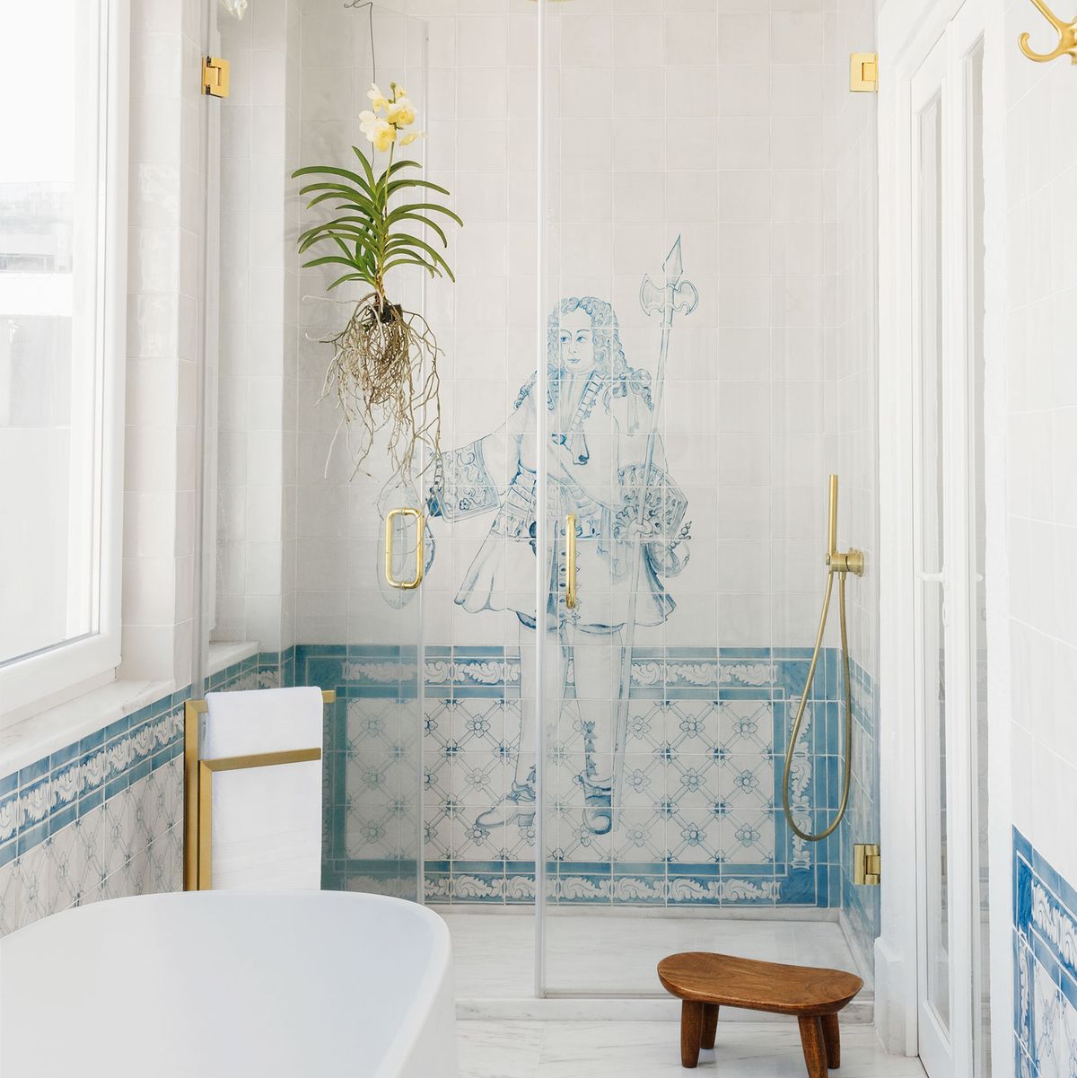 the main bathroom has a marble floor, deep bathtub, a small wooden stool, and in the glass doored shower, an image of an 18th century man with a staff and waistcoat is done in blue and white tiles