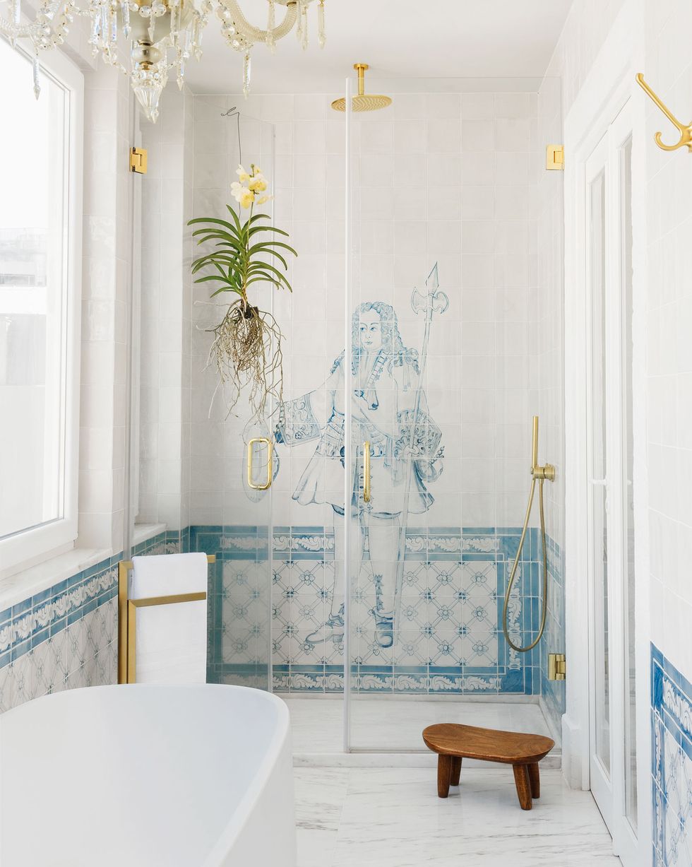 the main bathroom has a marble floor, deep bathtub, a small wooden stool, and in the glass doored shower, an image of an 18th century man with a staff and waistcoat is done in blue and white tiles