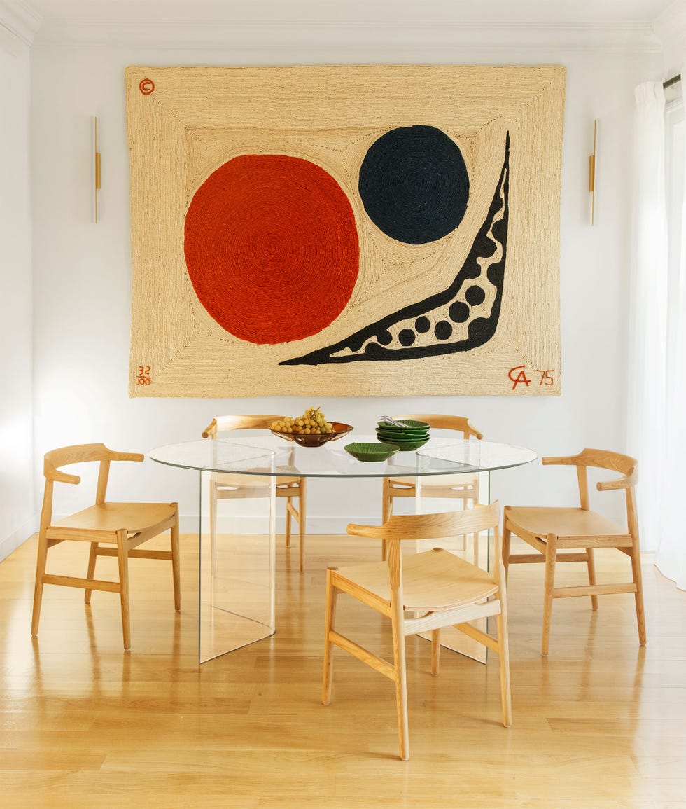 five simple wooden chairs surround a round glass dining table, the floor is wood, and on the wall is a large jute alexander calder tapestry with a red moon, a smaller black planet, and a boomerang shape