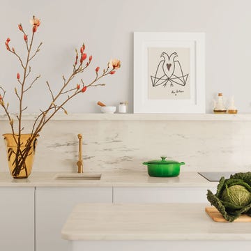 in the kitchen, an island and counter, with a built in sink, are topped with portuguese rosa marble, on the counter is a vase with tall salmon colored buds and a green saucepot, on a shelf is a drawing of two birds