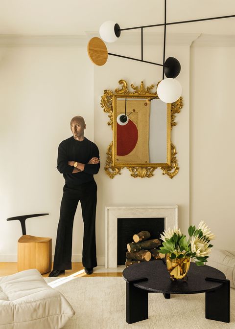 matthew harris, dressed in black, next to an antique gilded wall mirror, a low fireplace with logs, a modern wooden sculpted chair with a slender black metal back, and a light pendant with white and black orbs