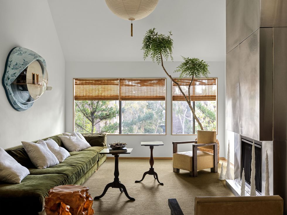 living room with high ceilings and a long sofa covered in a plant green velvet against the left wall and several small tripod tables scattered at center and opposite the sofa is a built in fireplace clad in metal and windows on the back wall with bamboo shades rolled up and a tall plant or tree peeking out from the corner of the room