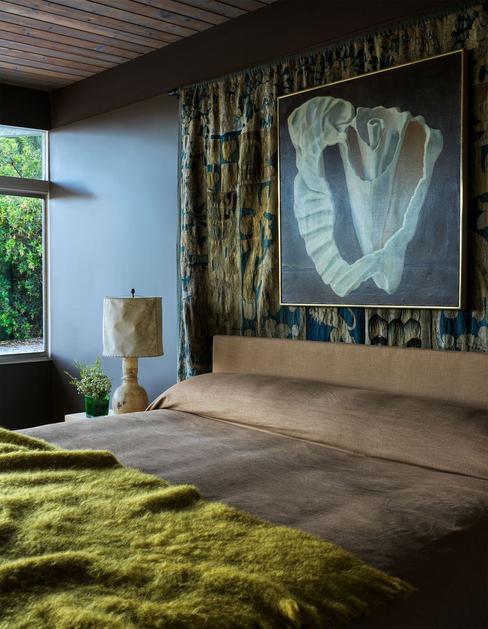 in a bedroom is a bed with a brown linen headboard and bedcover and a fuzzy green throw, a nightstand with a lamp is beside the bed, and behind the headboard hangs an antique tapestry and a surrealist painting