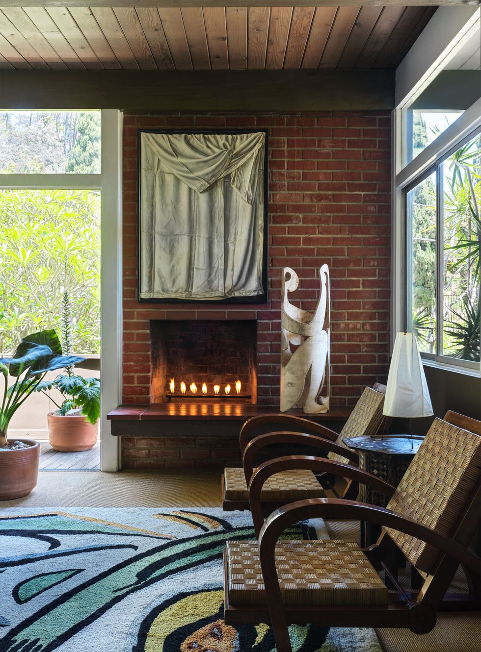 a family room has a brick wall with a fireplace and a painting, a standing paper sculpture, two vintage wood chairs with woven rush seats and backs, a colorful abstract rug, and a potted floor plant by a glass door