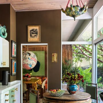 the kitchen has dark brown walls hung with small artworks, cabinets and drawers painted green and white, a small round antique wood table and two chairs with green leather seats, and a spanish majolica pendant
