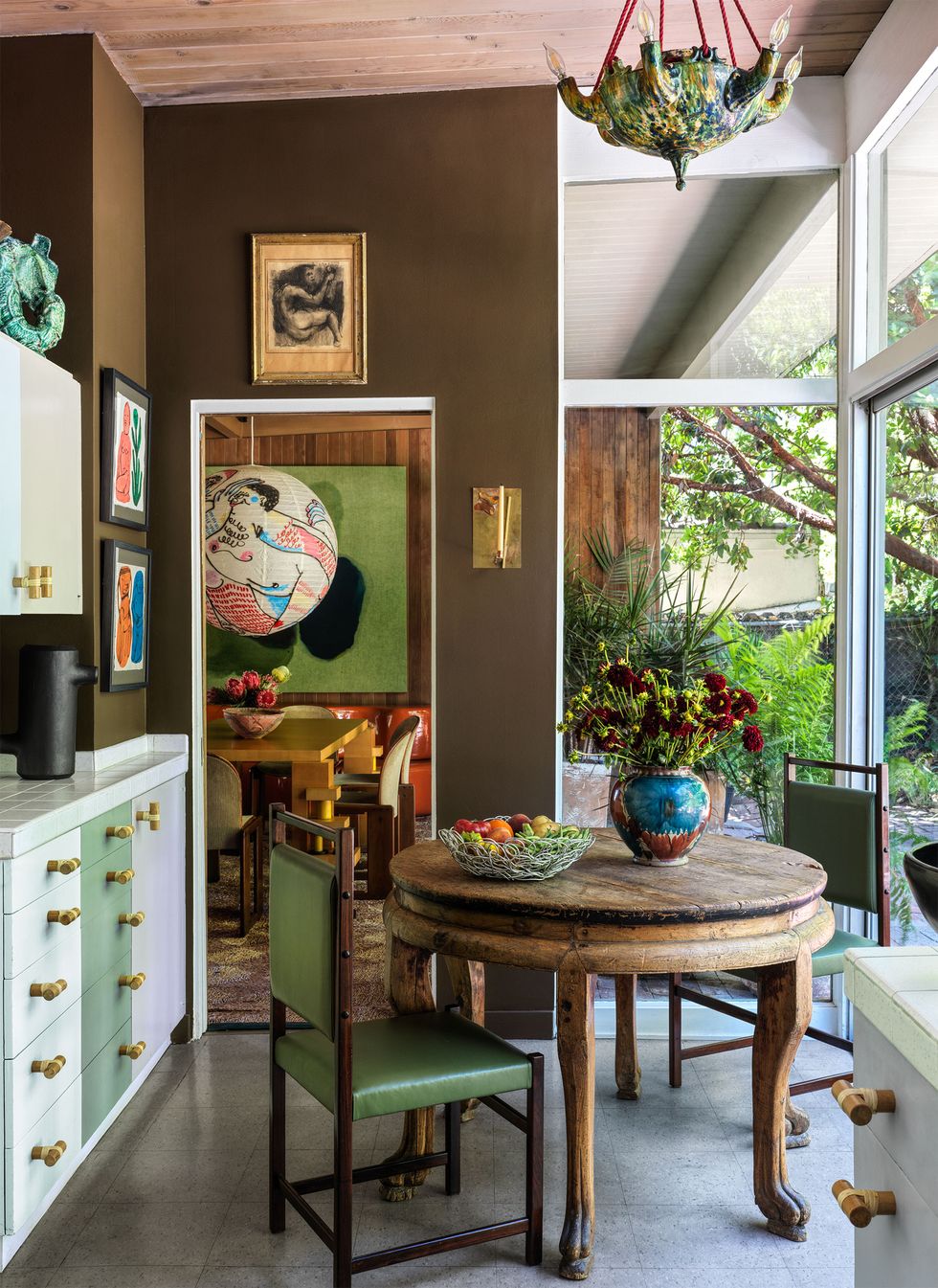 the kitchen has dark brown walls hung with small artworks, cabinets and drawers painted green and white, a small round antique wood table and two chairs with green leather seats, and a spanish majolica pendant