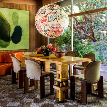 a dining room has wood paneled walls, a chunky wood table with six chairs, a glass wall with door opening to an outside terrace, a large handpainted globe pendant, and a large abstract painting with green hues and shapes