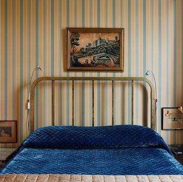 a guest bedroom has striped wallpaper, a metal bed headboard and deep blue velvet bedcovering, vintage tables on each side hold vintage lamps with orange shades, a painting is above the headboard