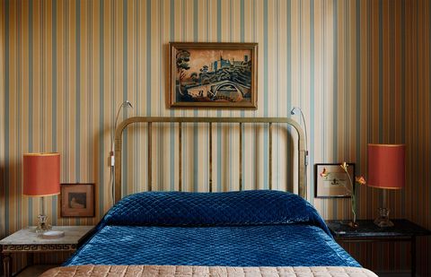 a guest bedroom has striped wallpaper, a metal bed headboard and deep blue velvet bedcovering, vintage tables on each side hold vintage lamps with orange shades, a painting is above the headboard