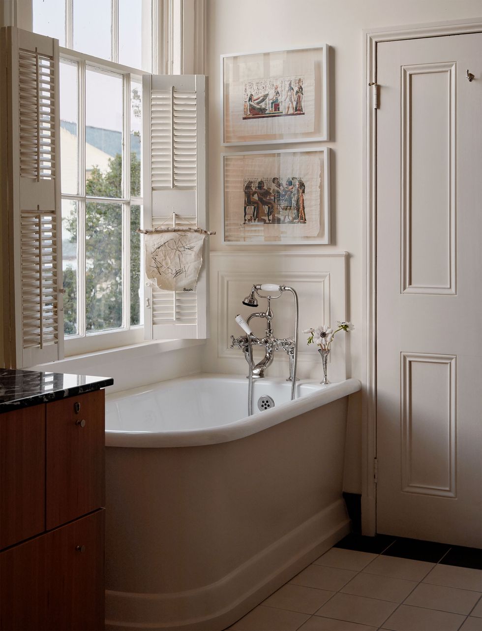 a deep tub sits in the corner of the main bathroom next to the door and under a window outfitted with interior shutters, above fixtures are paintings on silk and next to the tub is a vanity with a black marble top