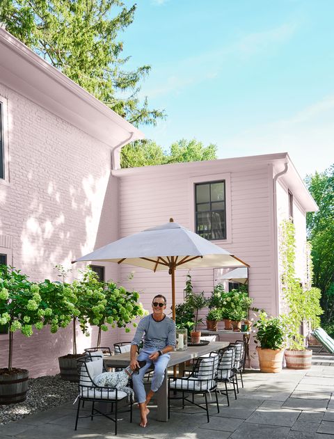 on a slate patio with flowering shrubs is a man sitting on a teak table with eight metal chairs with cushions, a dog seated on one, and a sun umbrella next to a brick and clapboard house painted light pink