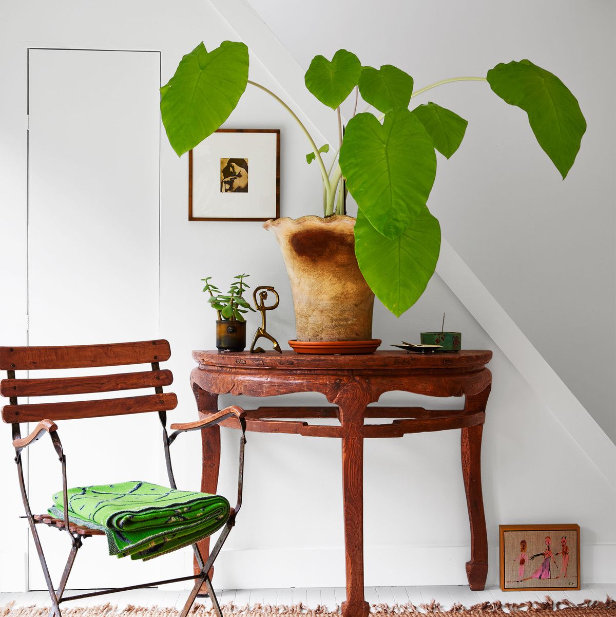 in the entry to a cottage is a bistro style chair with a green folded towel on the seat, a demilune table with a large potted plant and a small brass sculpture, a sisal rug, an artwork on the wall and one on the floor