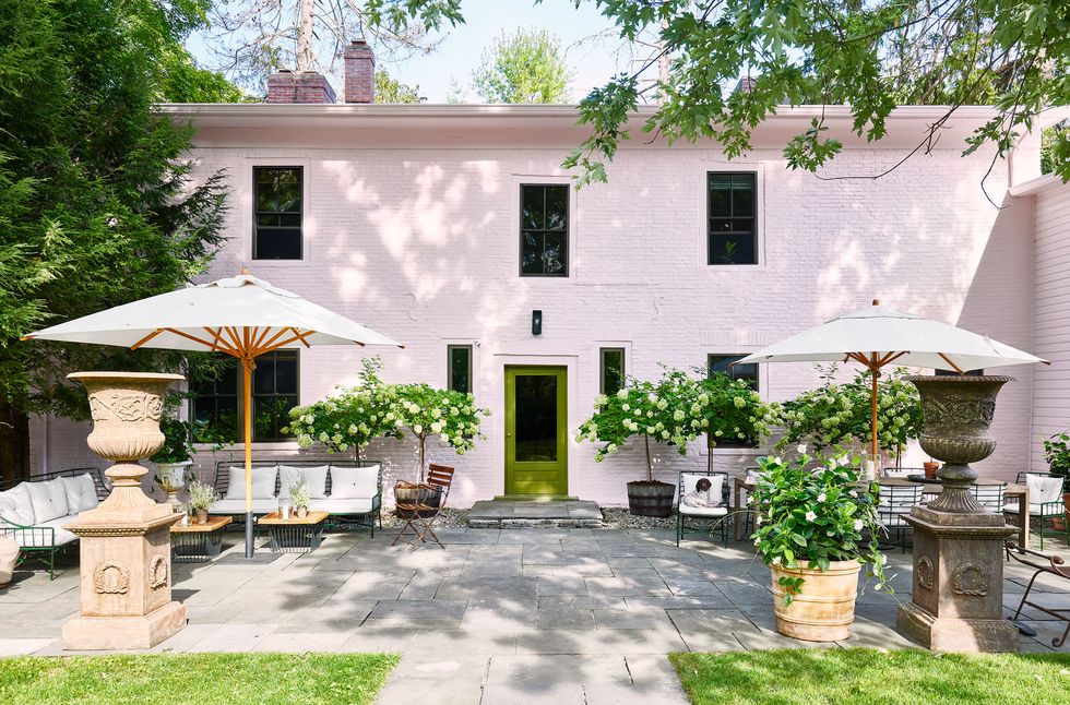 the terrace of a pink two story house has stone flooring, a seating area with sofas and an umbrella at left and a dining table and chairs and umbrella at right, terra cotta urns, and a green door to the house