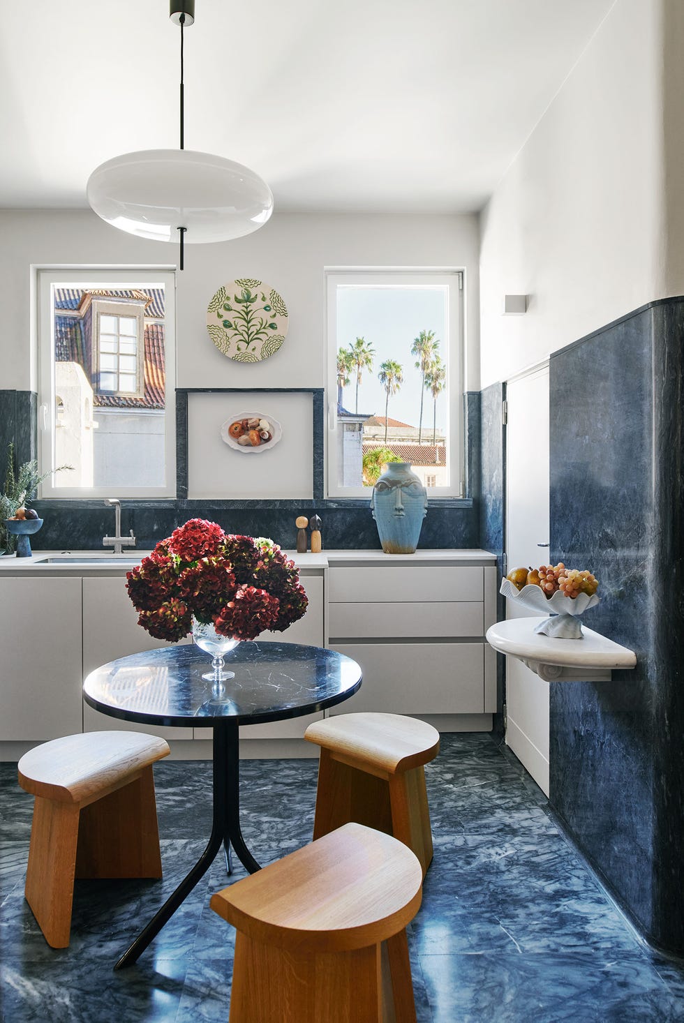 Elad Yifrach - ELLE DECOR a kitchen floor, backsplash, partial wall, and a round table are a blue marble, three stools are oak, two windows are above white cabinets and sink, a floating shelf has a fruit bowl, a spheroid pendant above table