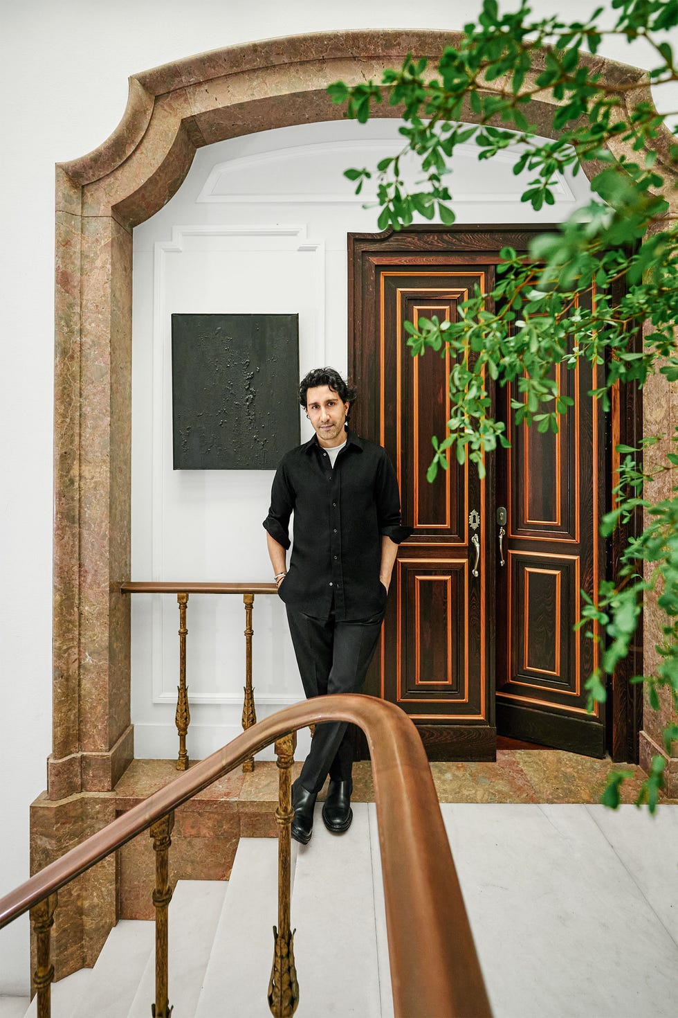 the owner in a black shirt and pants stands in an arched entrance at the top of a marble staircase with copper balustrades and railings near the double doors of his apartment