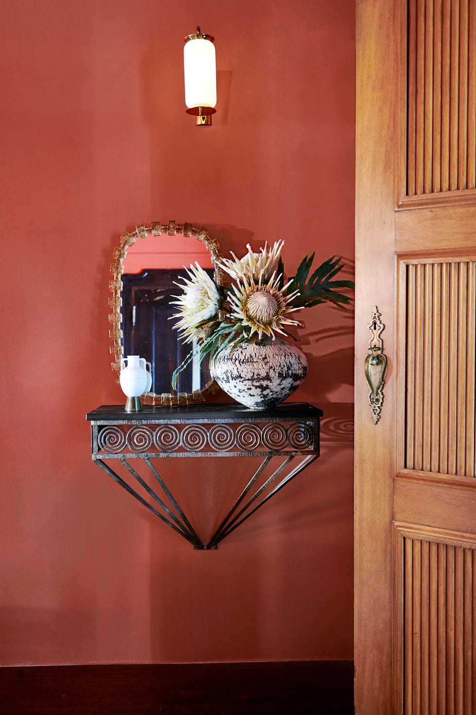 a carved wood door with brass hardware opens to an entry hall painted in a red with orange undertones, a decorative iron shelf holds vases and an antique mirror, a glass and brass sconce is above shelf