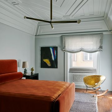 in a guest room is a bed with persimmon colored velvet headboard, bolster, and cover, a striped berber rug, a nightstand, a woven brass rocking chair, and abstract artwork, and a three armed pendant light