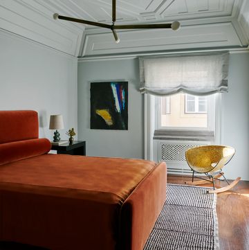 in a guest room is a bed with persimmon colored velvet headboard, bolster, and cover, a striped berber rug, a nightstand, a woven brass rocking chair, and abstract artwork, and a three armed pendant light