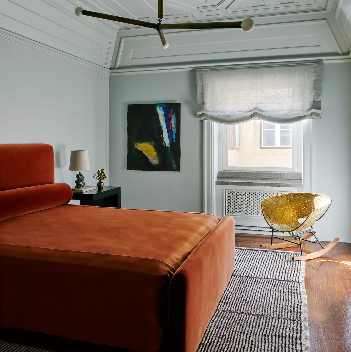 in a guest room is a bed with persimmon colored velvet headboard, bolster, and cover, a striped berber rug, a nightstand, a woven brass rocking chair, and abstract artwork, and a three armed pendant light