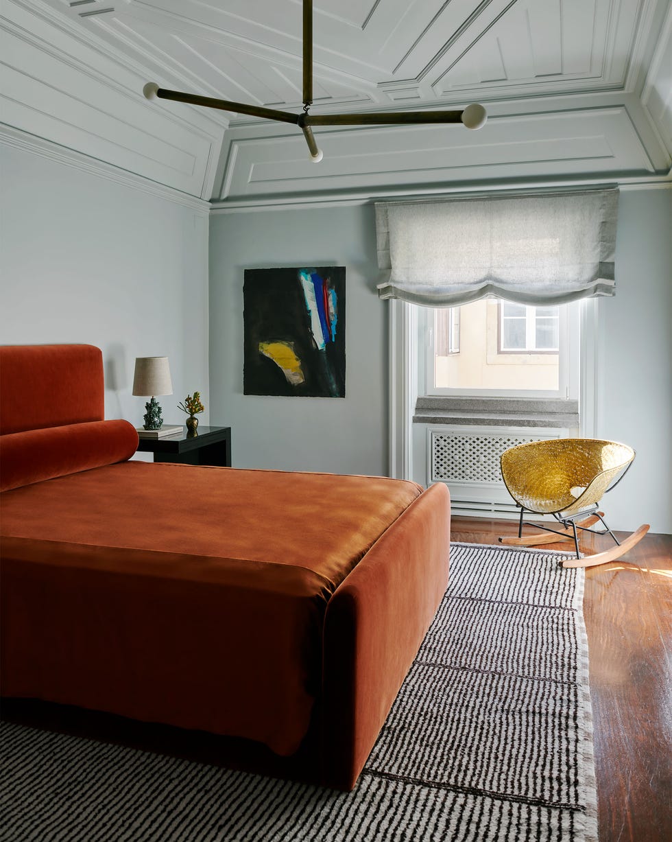 in a guest room is a bed with persimmon colored velvet headboard, bolster, and cover, a striped berber rug, a nightstand, a woven brass rocking chair, and abstract artwork, and a three armed pendant light