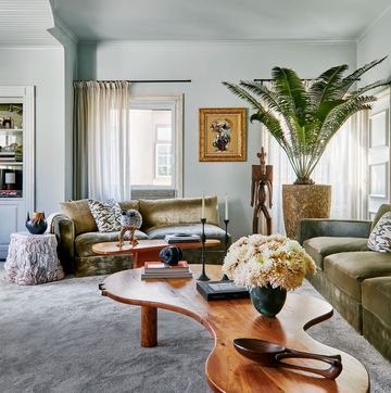 a living room with two sofas in moss green velvet with decorative pillows, a small and larger cocktail table with curvy edges, a terra cotta side table, female fertility sculpture, tall palm plant, light gray carpet