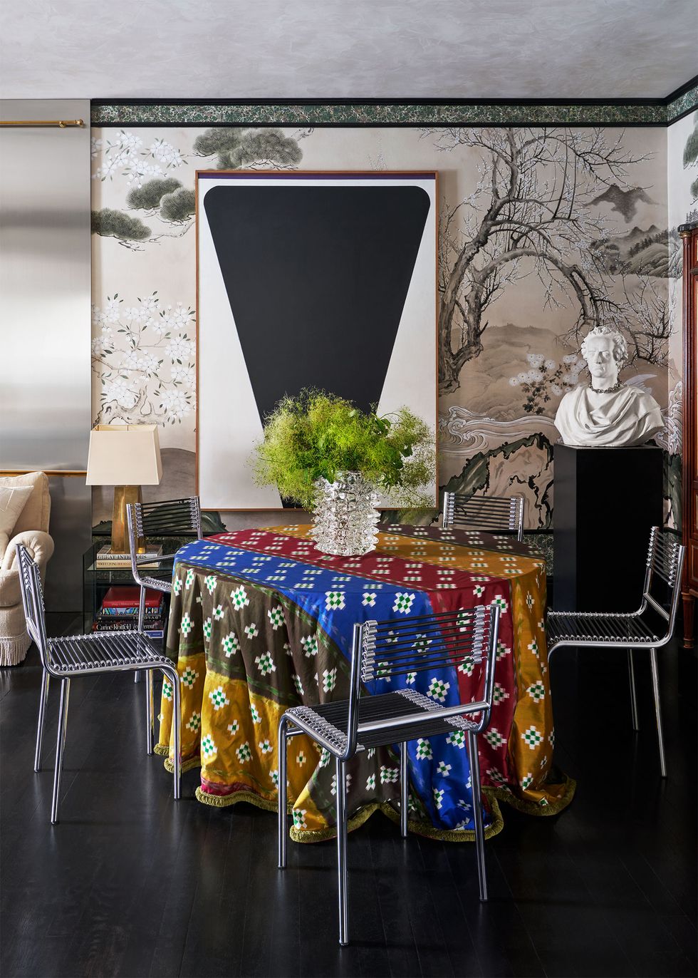 a round dining table covered with panels of brightly colored panels, four steel chairs, a glass vase with greens, a bust sculpture on a black plinth, a framed artwork of a black upside down triangle