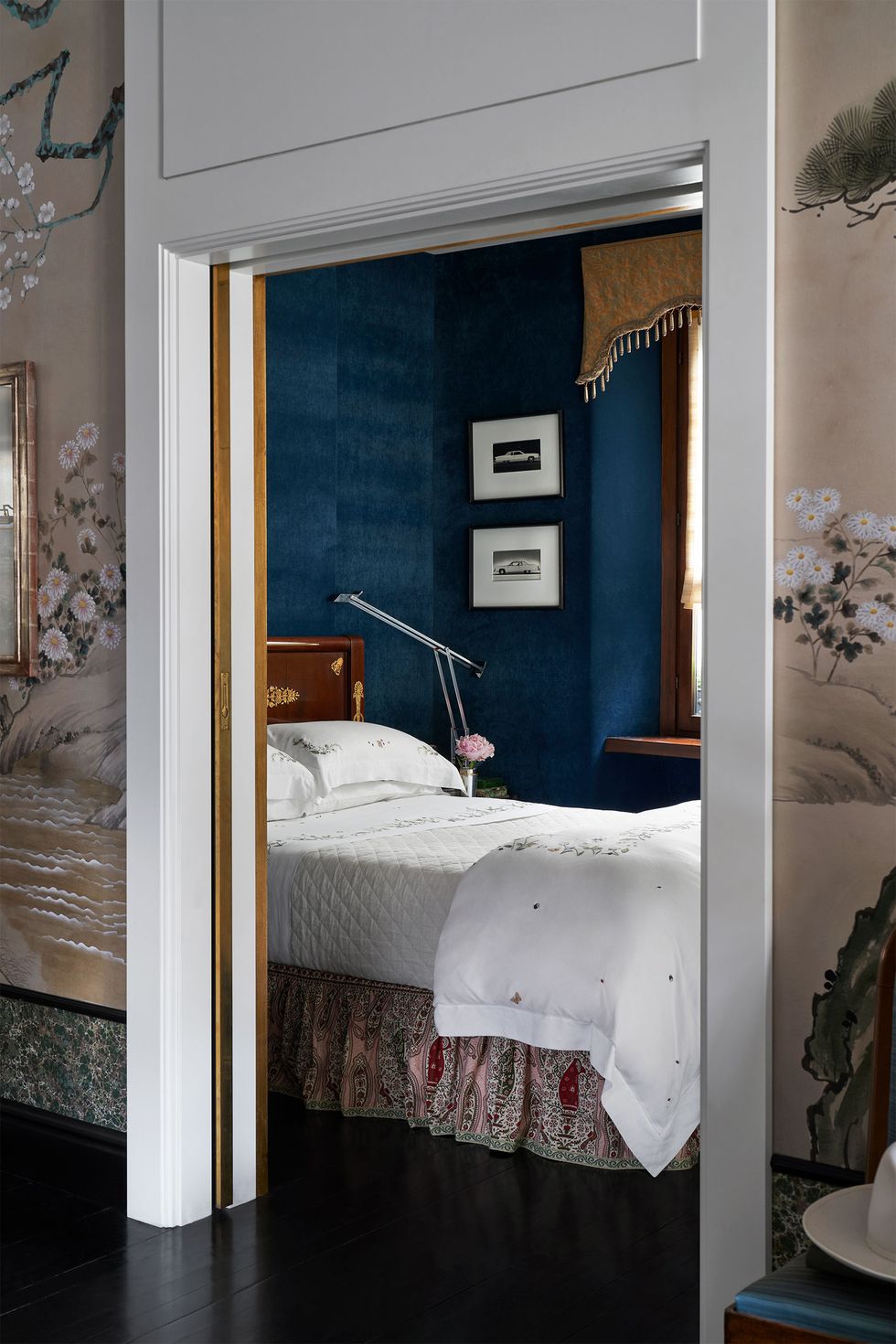 a bedroom, seen from a hallway, has deep blue velvet walls, a window valance with short tassels, two small framed artworks, a bed with a wood headboard, white linens, and printed bedskirt, floor lamp beside bed