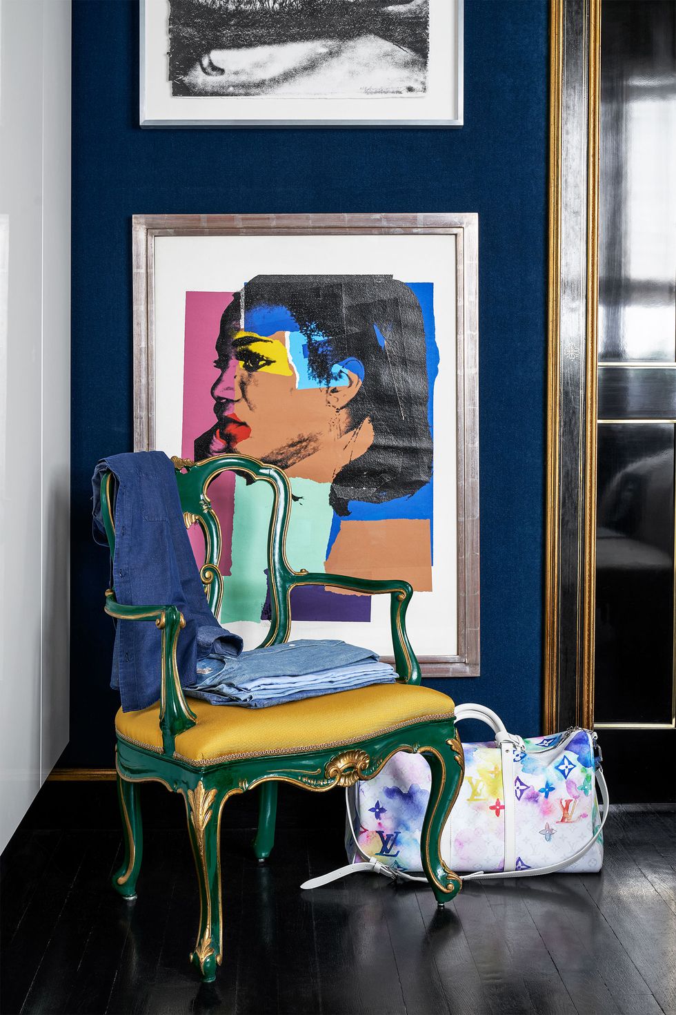 in a bedroom corner is a vintage chair with a green curvy frame and gold fabric seat with folded clothing , a colorful weekend bag sits on a dark wood floor, blue velvet walls with a collage of a woman's face