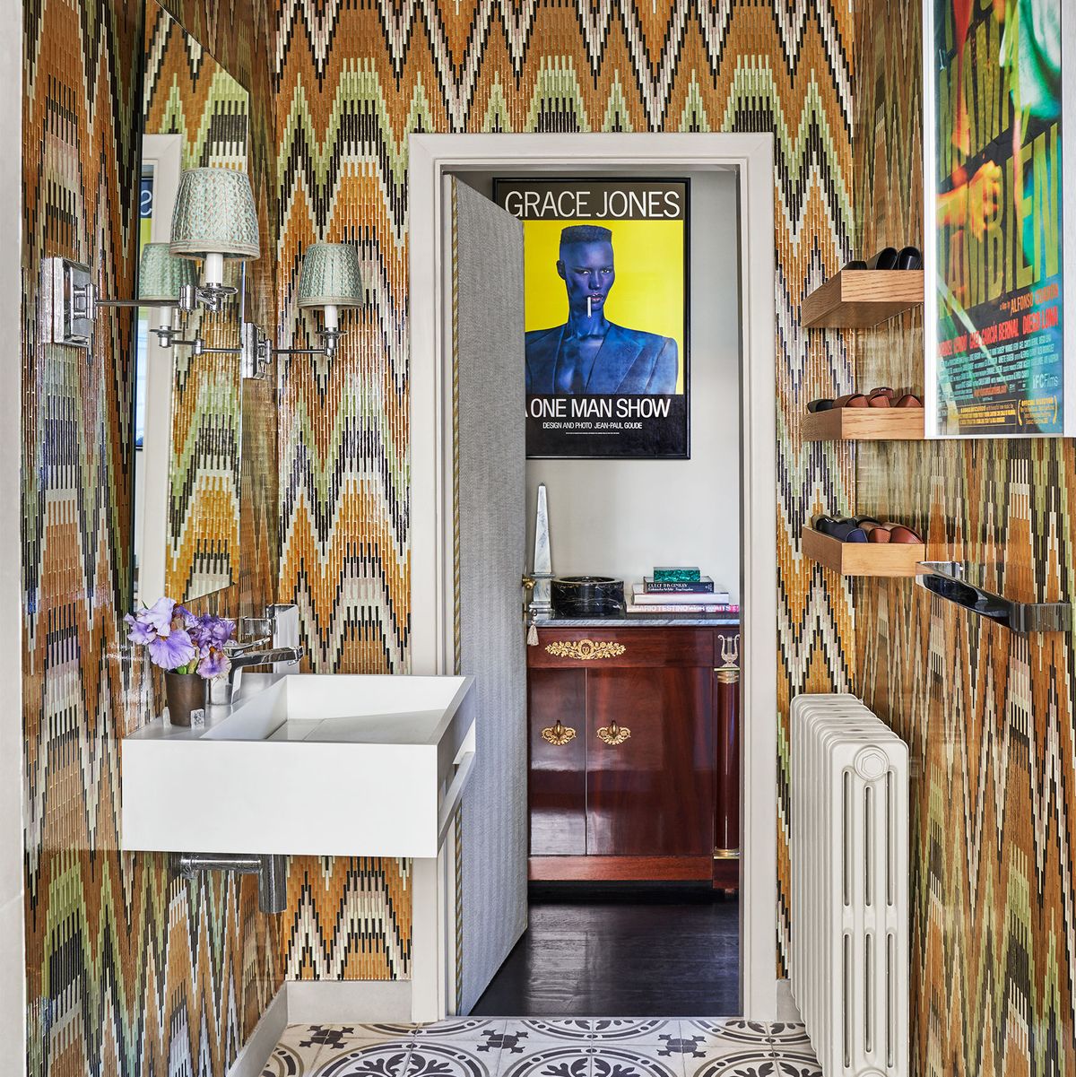 a washroom with bright zigzaggy wallpaper, sink on one wall and radiator on the facing wall, moroccan floor tiles, a door opens to a room with a poster of grace jones hanging above a wood vanity