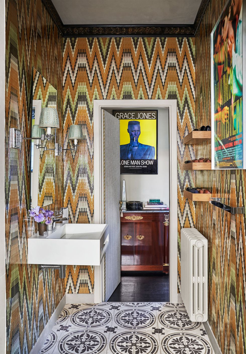 a washroom with bright zigzaggy wallpaper, sink on one wall and radiator on the facing wall, moroccan floor tiles, a door opens to a room with a poster of grace jones hanging above a wood vanity