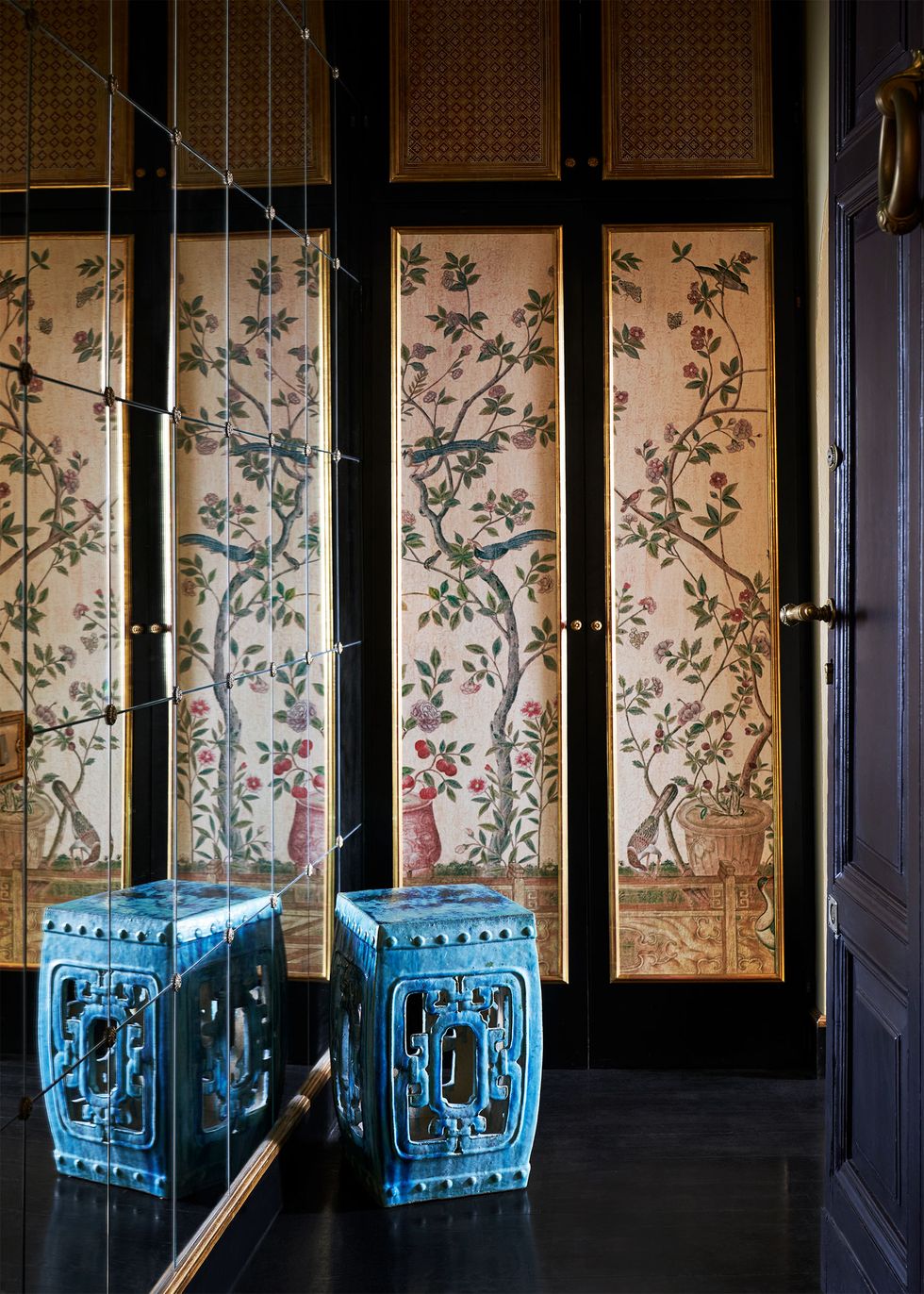 an entrance hall has a mirrored wall on one side and a far wall of closet doors with panels covered in a flowered fabric, blue chinese stool, dark wood floor