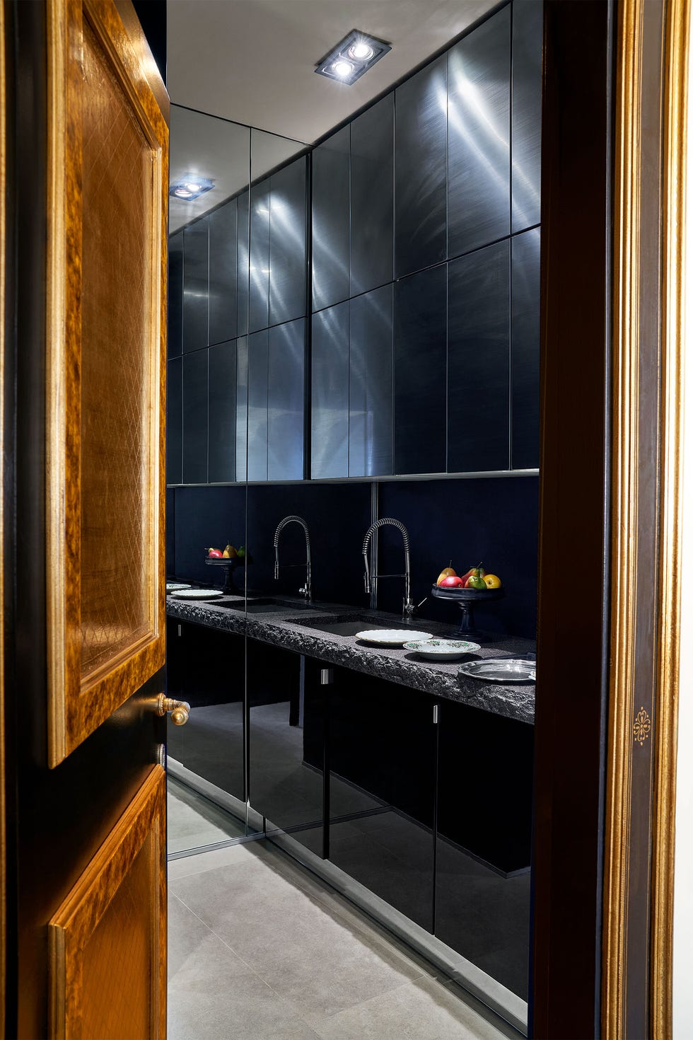 Eric Egan - ELLE DECOR a door opens to a kitchen with light stone flooring, black cabinets above and below sink, dark gray stone countertop, three plates and a fruit bowl on counter