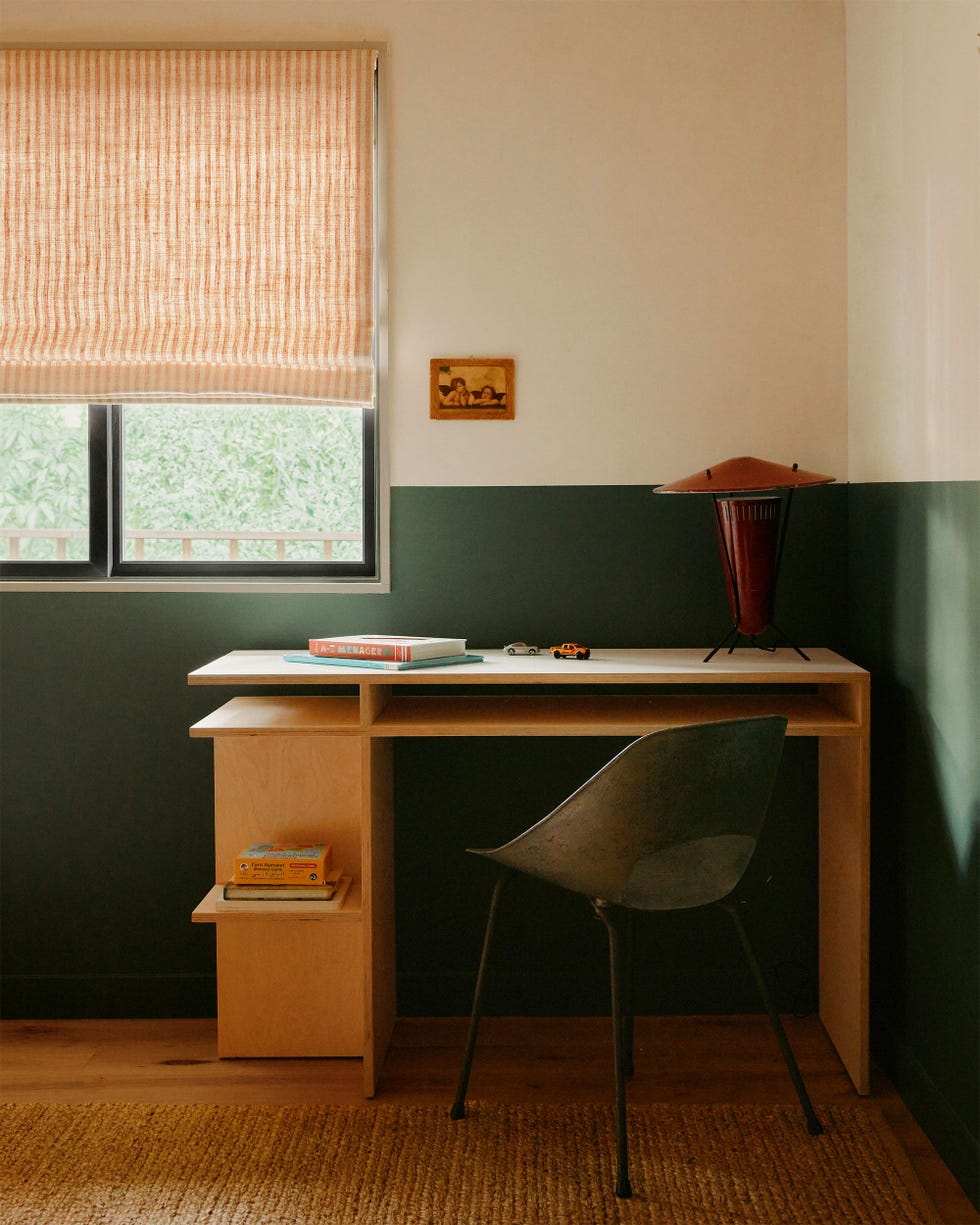 a modern wood desk with a lamp and a chair in a room in muted colors of cream and green with a window with a striped shade