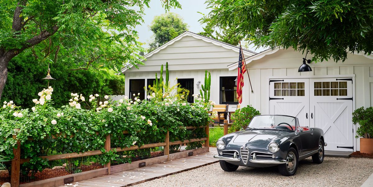 an alfa romeo spider convertible in a gravel driveway in front of a white garage with manual doors, a flag, walkway next to a fence with flowering rose bushes, white ranch house with windows, mature trees
