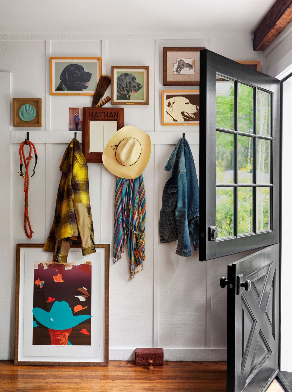 an entry with a black dutch door, wood floor, white walls with hooks holding jackets, scarf, and hat, a mirror, several framed pictures of dogs, a larger framed image of a man in a hat leans against the wall