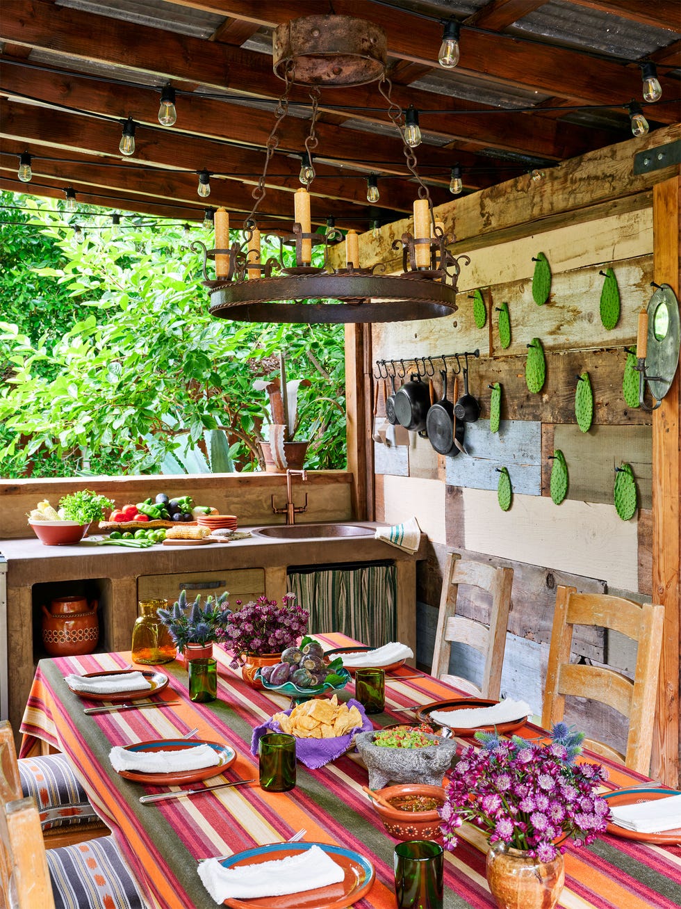 Nathan Turner and Eric Hughes - ELLE DECOR outdoor kitchen with cast iron skillets hanging on rustic wood wall, sink and counter with produce on top and cabinets below, set dining table with bright striped cloth and flowers, wood chairs with patterned seat cushions