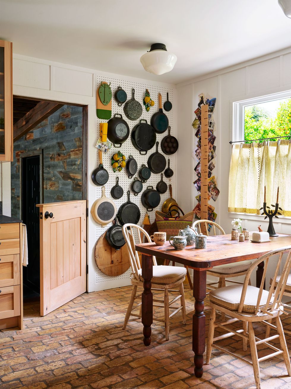 kitchen with old brick flooring, dutch door open at top, pegboard covering one wall with pots hanging from top to bottom, wood table with wood chairs and fabric seat cushions, window with light yellow half curtain