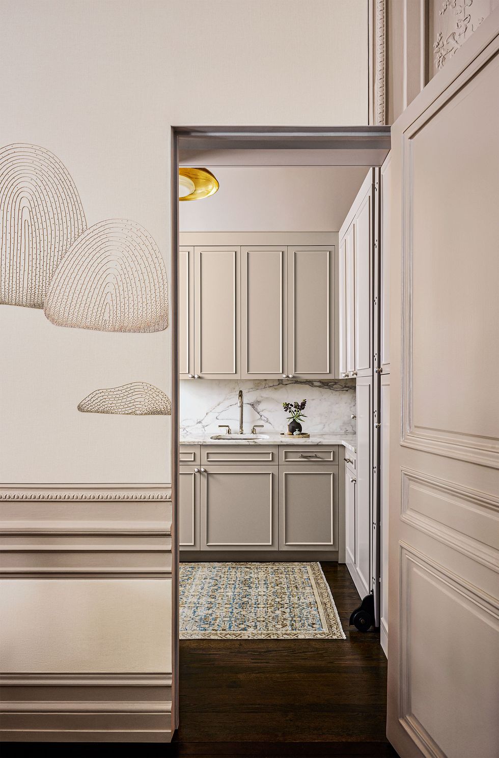 a wall panel with a cloud patterned wallcovering next to a door leading to a kitchen with taupe colored cabinets and with a sink and white marble backsplash and counters, a subtle patterned rug sits on dark wood floor