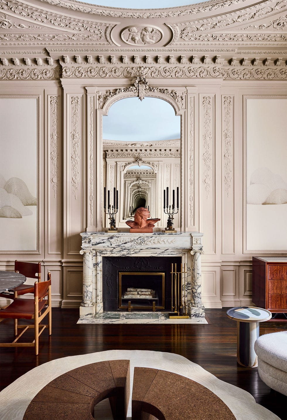 Jean Liu - ELLE DECOR a fireplace with a marble surround and mantel with a sculptured bust atop it and a mirror above, decorative molding on walls and ceiling, light amorphous shaped rug, two half moon cocktail tables