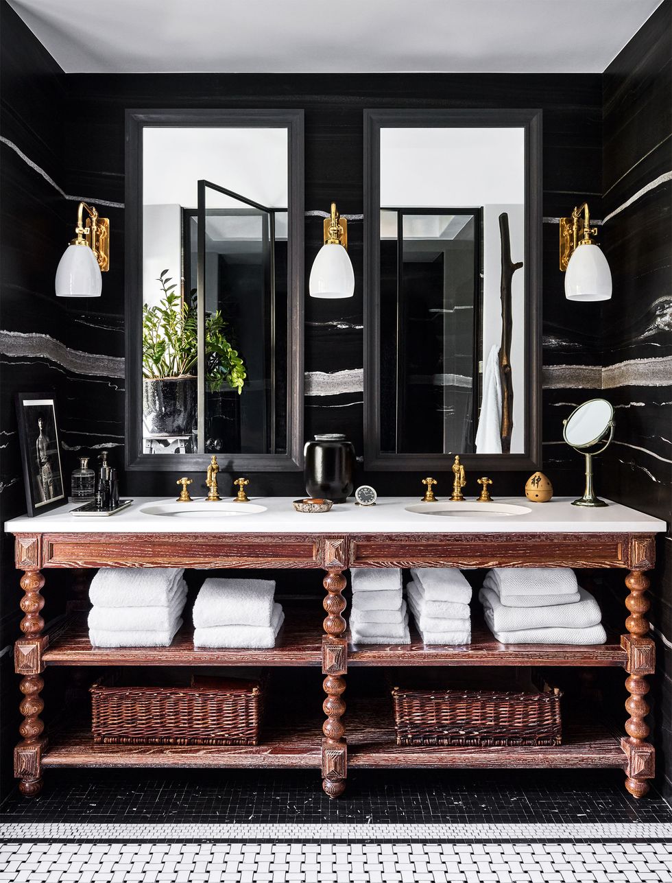 a bathroom with black walls, three sconces flanking two mirrors, wooden vanity with turned legs and two shelves holding towels and baskets, a white marble top with two small round sinks with brass fittings