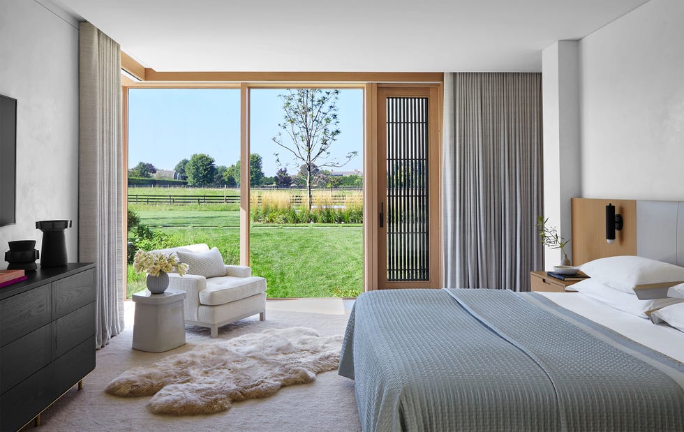 a bedroom with curtained floor to ceiling windows looking out to grassy fields, bed with gray headboard and coverlet, attached nightstand with small sconce, , dresser, armchair, sheepskin rug on top of light carpet