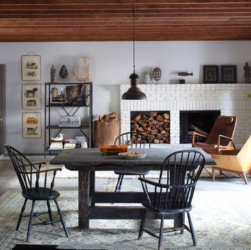wood beam ceiling living dining space with a white brick fireplace at far end and a wood insert next to it and some modern wood club chairs and a dark square farmhouse table and chairs in the foreground and a kitchen set up to the left