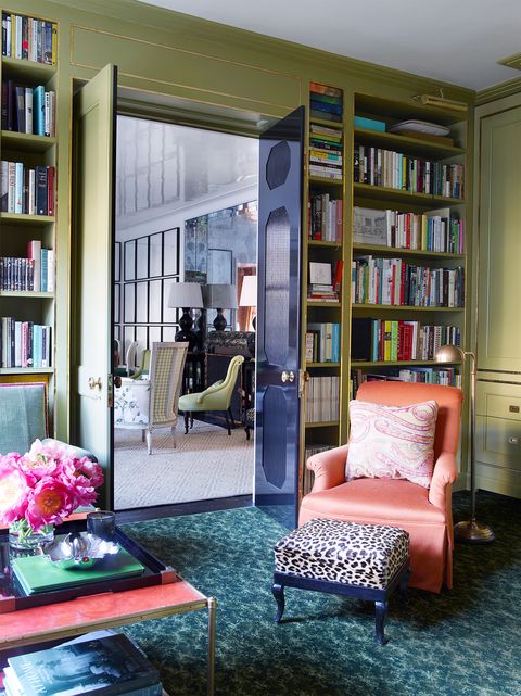 library with floor to ceiling shelving and aqua blue patterned carpet and a coral slipper chair and small stool at center with a view through a door into the living area
