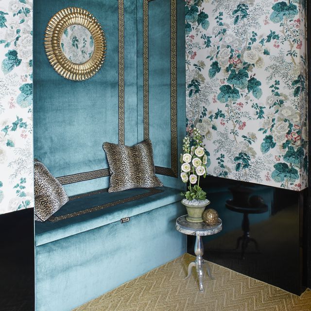 a room designed by young huh for the 2014 kips bay decorator show house it is a corner with a blue velvet banquette withe silver green key detail and silver and black pillows in a sort of tiger pattern and a gold chevron rug and a floral wallpaper on the wall
