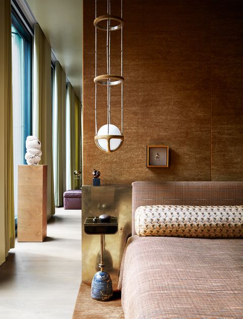 a brass bed with a fabric headboard and matching bedcover, the wall behind covered in an amber gold fabric, a marble and leather table next to the bed, and a brass, glass, and suede pendant light above