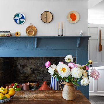 weathered dark wood kitchen island with a vase of peonies and a bowl of lemons, stone fireplace with blue painted wood surround, objets d’art on mantle and three plates on wall above