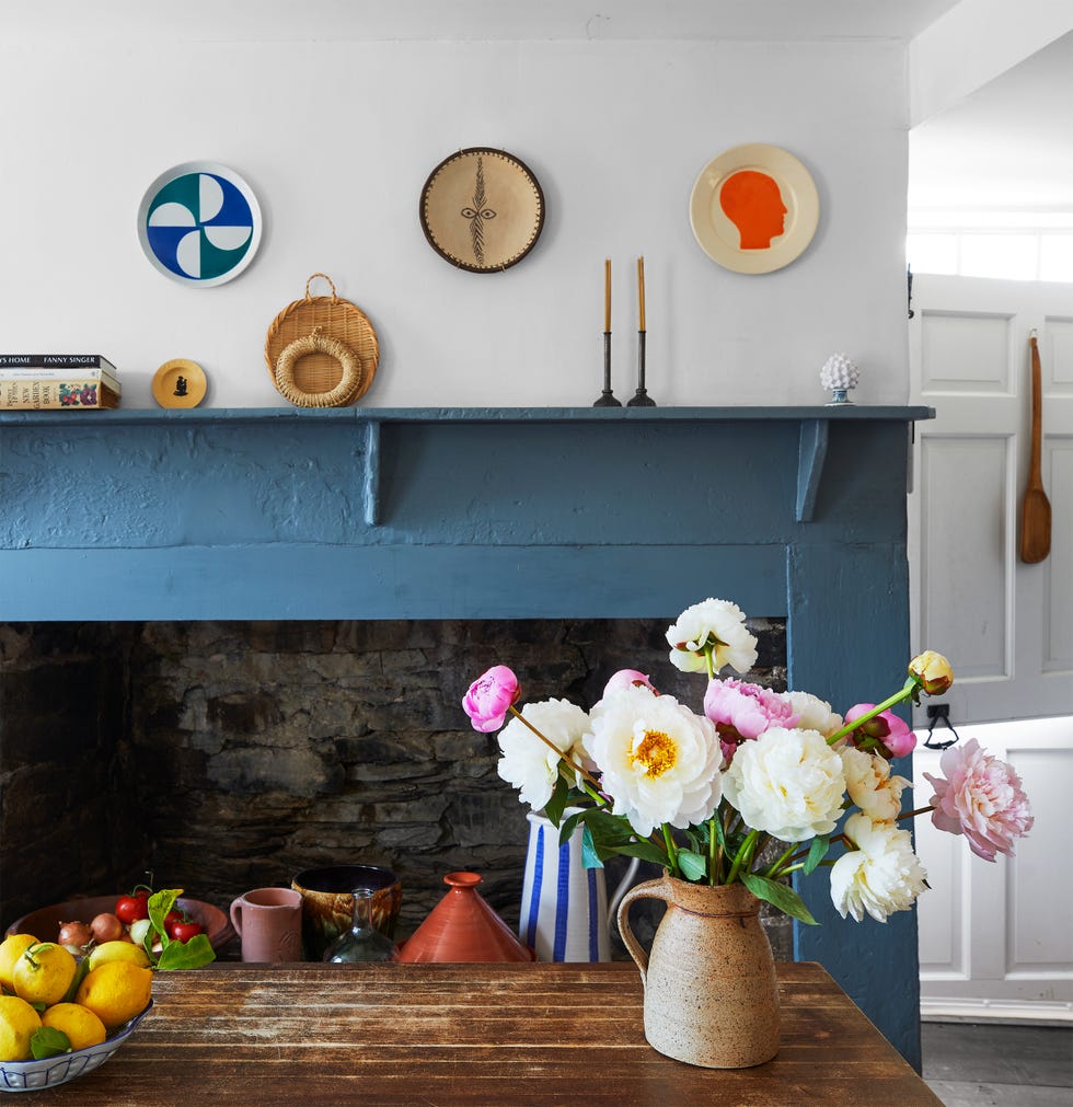 Archer-Coite - ELLE DECOR weathered dark wood kitchen island with a vase of peonies and a bowl of lemons, stone fireplace with blue painted wood surround, objets d’art on mantle and three plates on wall above