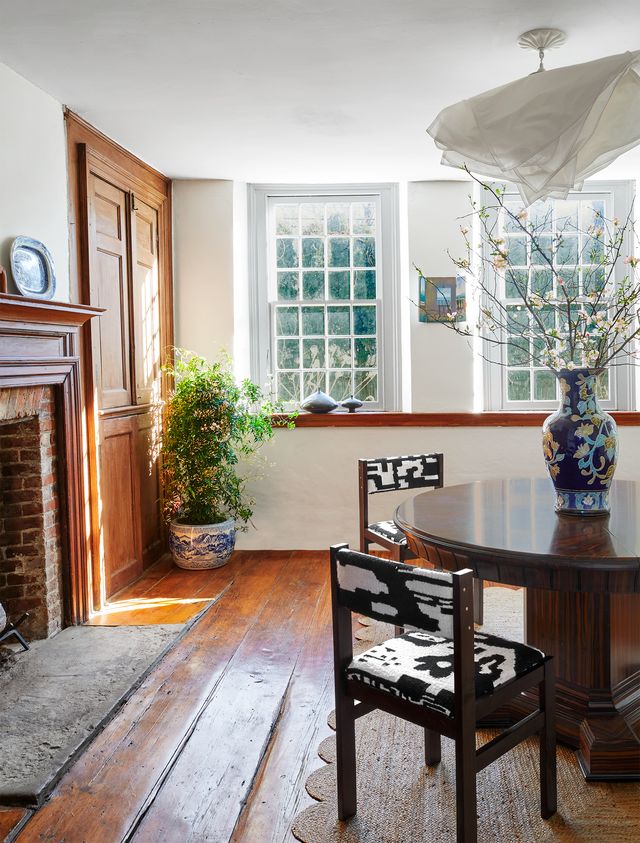 a dark wood vintage dining table and two chairs with fabric seats and backs on a jute rug atop worn wide plank wood flooring, a brick fireplace with wood surround, and a floor plant in a vintage japanese pot